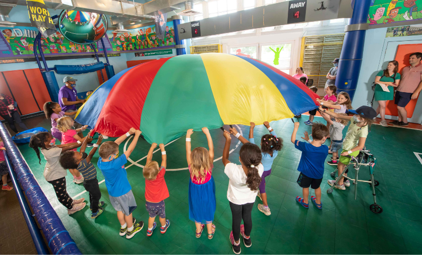 A group of children working together to hold and lift up a giant colorful parachute during an interactive program for kids at Port Discovery