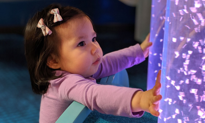 Toddler looking at and touching lighted bubble tubes inside of a sensory-friendly toddler play area