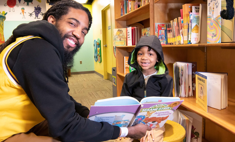 An adult and child smiling at the camera as they read a book together in one of Port Discovery's play areas