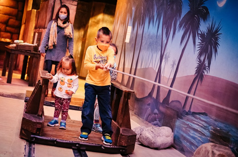 Two children standing on a wooden raft as one uses a rope to pull their way across a pretend river