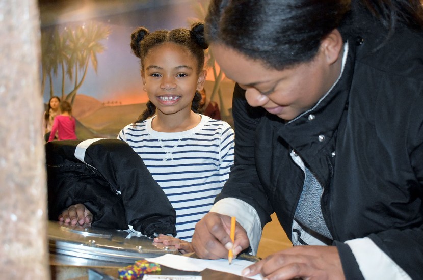 A child smiling at the camera as an adult writes on a piece of paper as part of an activity featured in Port Discovery's Adventure Expeditions exhibit