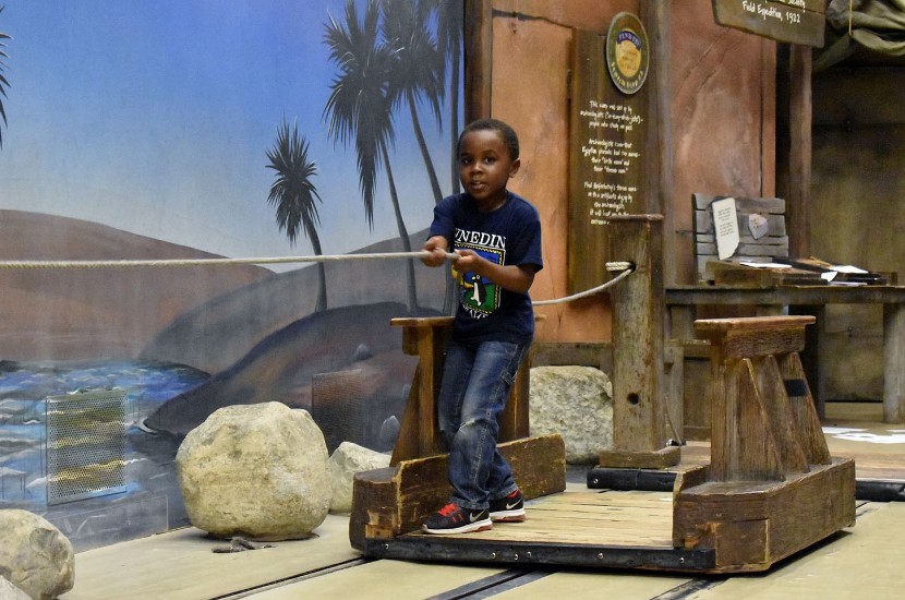 A child pulling themselves across a pretend river on a wooden raft featured in an exhibit exploring Ancient Egypt