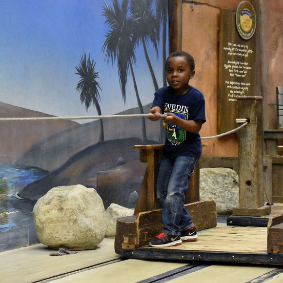 A child pulling themselves across a pretend river on a wooden raft featured in an exhibit exploring Ancient Egypt
