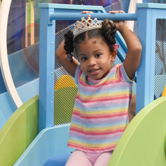 A toddler wearing a tiara getting ready to slide downa miniature blue and green slide featured in one of Port Discovery's toddler play areas