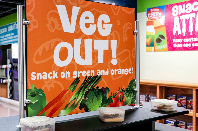 A view of a pretend convenience store featuring a sign reading "Veg Out! Snack on green and orange" featured in one of Port Discovery's play areas for children