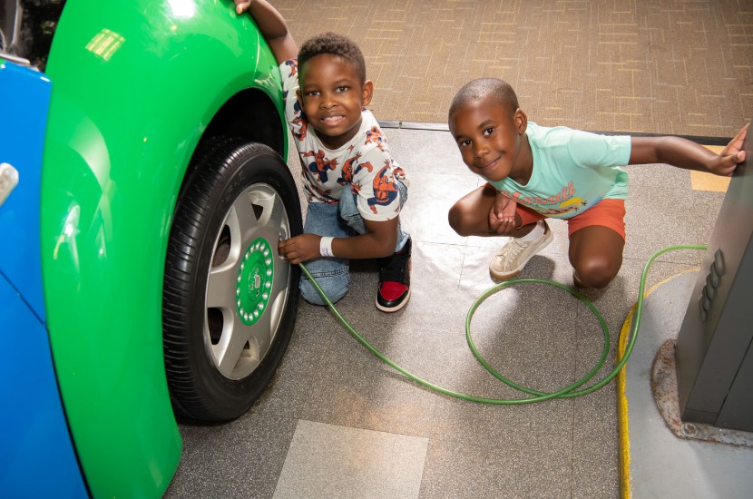 Two young children kneeling and smiling at camera as they use an air hose to fill up the tires of a colorful car