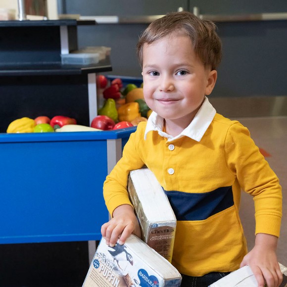 A child smiling and carrying several cereal boxes as they explore Port Discovery's pretend convenience store