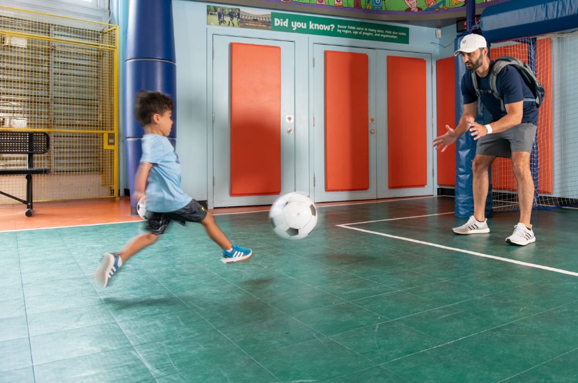 A boy preparing to kick a soccer ball at an adult standing in the goal area of a pretend indoor sports field