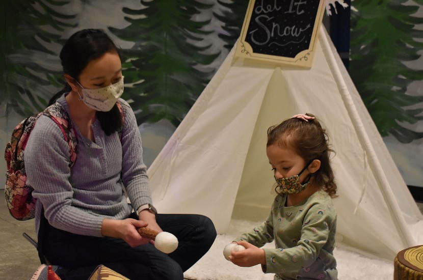 An adult and a young child sitting on the floor and pretending to roast marshmallows near a white tent surrounded by painted trees