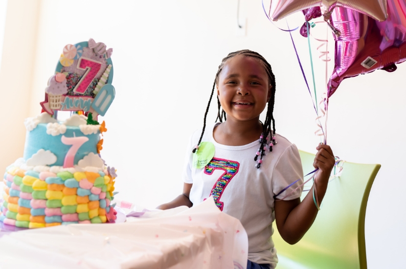 A child holding pink and purple balloons and sitting by a birthday cake