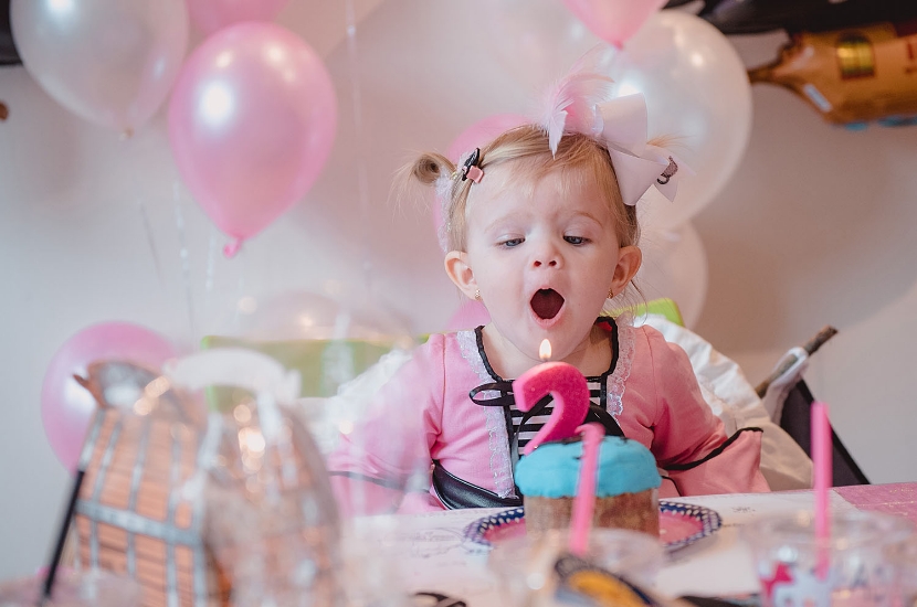 A young girl surrounded by balloons prepares to blow out a candle on a small birthday cake
