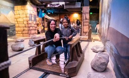An adult and a child standing on a wooden raft work together to pull themselves across a pretend river featured in Port Discovery's interactive Adventure Expedition exhibit