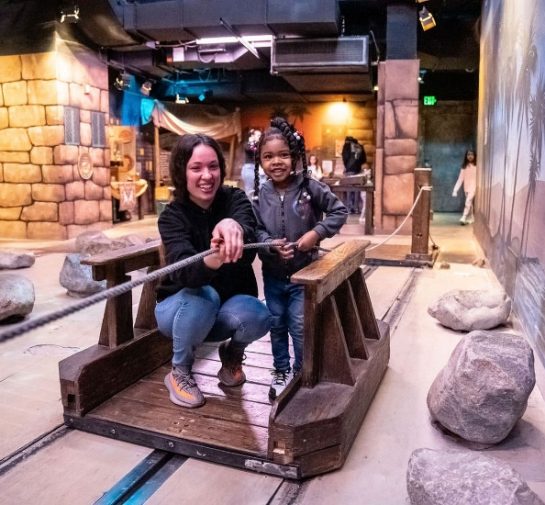 An adult and a child standing on a wooden raft work together to pull themselves across a pretend river featured in Port Discovery's interactive Adventure Expedition exhibit