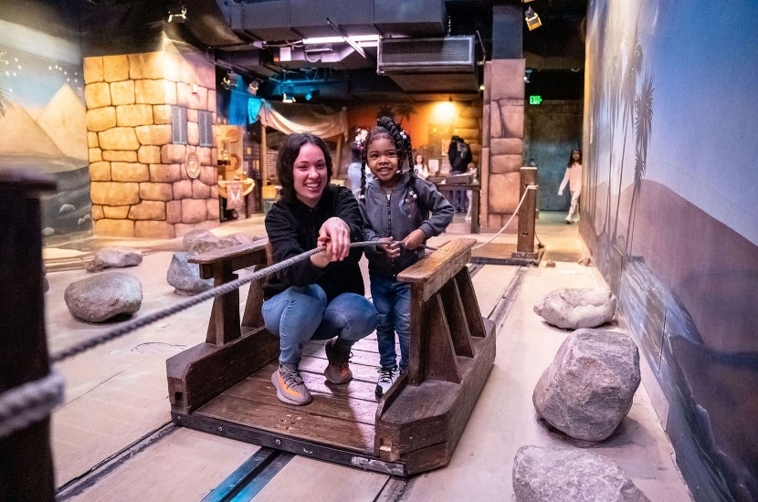 An adult and a child standing on a wooden raft work together to pull themselves across a pretend river featured in Port Discovery's interactive Adventure Expedition exhibit