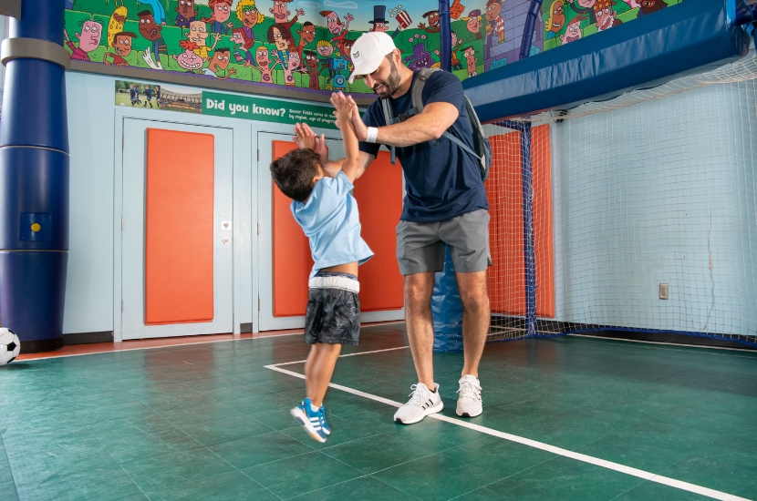 A child jumps up to give an adult a high five while they play in an indoor pretend sports field at Port Discovery