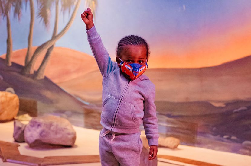 A young child standing on a rock with one hand lifted towards the sky as they celebrate making their way across a pretend river in Port Discovery's Adventure Expeditions exhibit