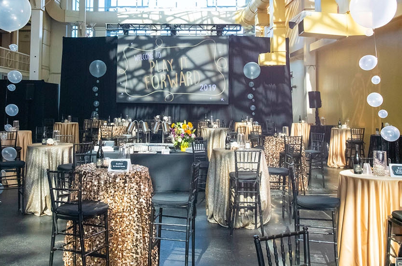 Rows of decorated tables and chairs and a large screen setup in a long, bright atrium that hosts private events at Port Discovery