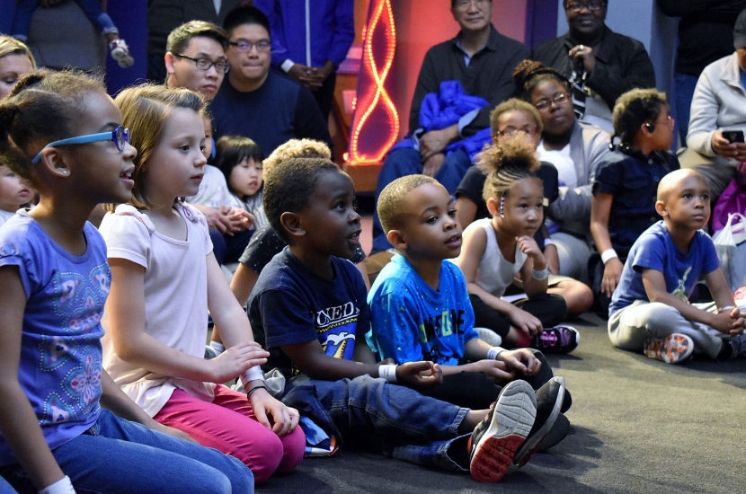 A large group of children and adults sit on the floor as they watch and participate in a group program at Port Discovery
