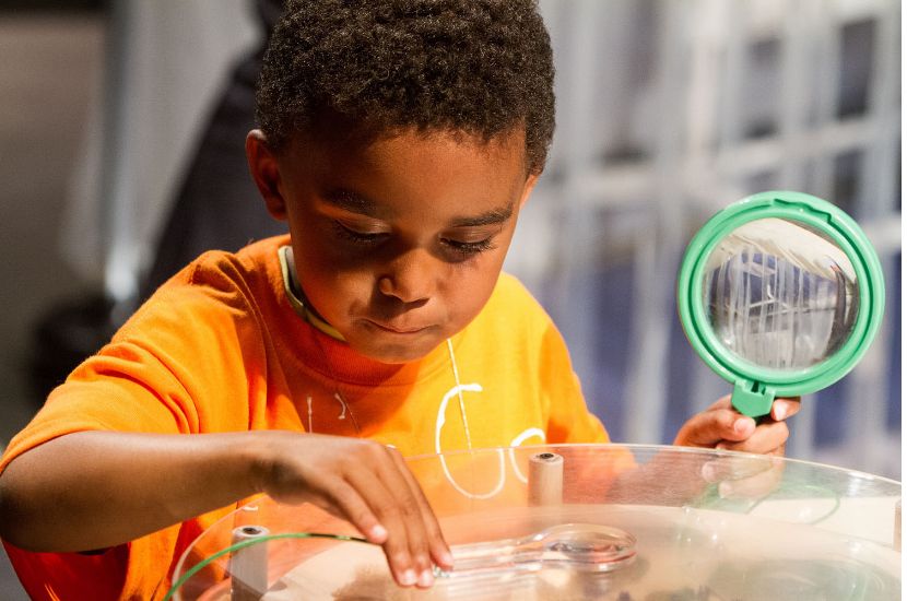 A child holds a magnifying glass while inspecting and playing with an interactive exhibit piece