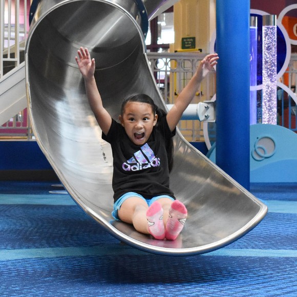 Excited girl with arms raised and big smile after landing at the bottom of Port Discovery's giant metal slide