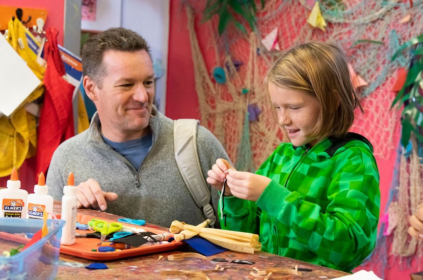An adult looking on as a child uses a variety of supplies to create and decorate an art project at Port Discovery