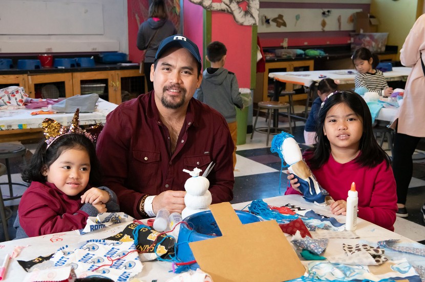 An adult surrounded by two children sitting at a table filled with art and craft supplies in Port Discovery's interactive art studio