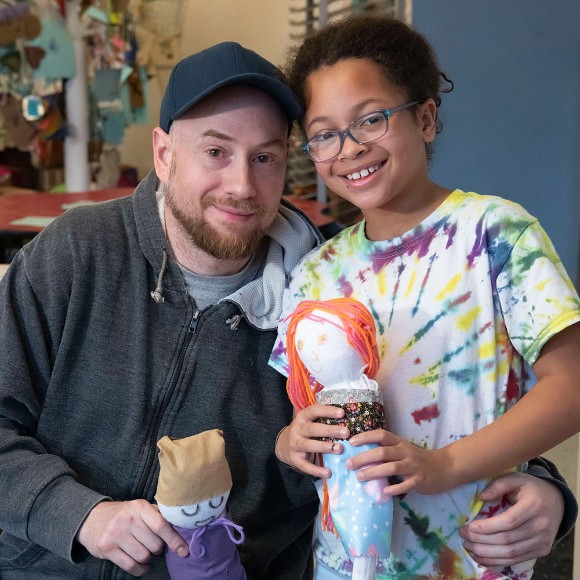 A man and a child smiling as they hold up sock puppets they created together in Port Discovery's art studio