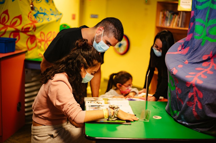 Two adults in masks reading a book while another child and adult play in one of Port Discovery's indoor play areas for children