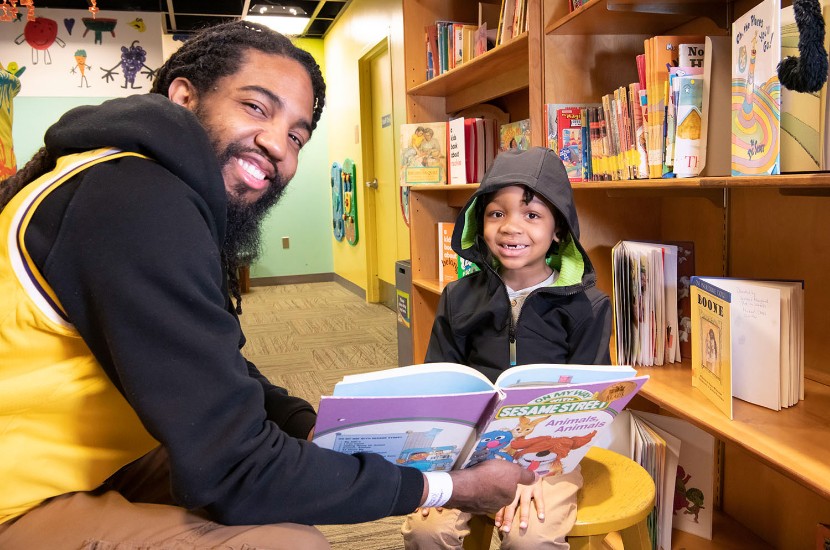 An adult and child smiling at the camera as they read a book together in one of Port Discovery's play areas