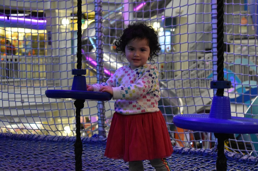 Young child standing on mesh climbing play area featuring blue seats held up by ropes