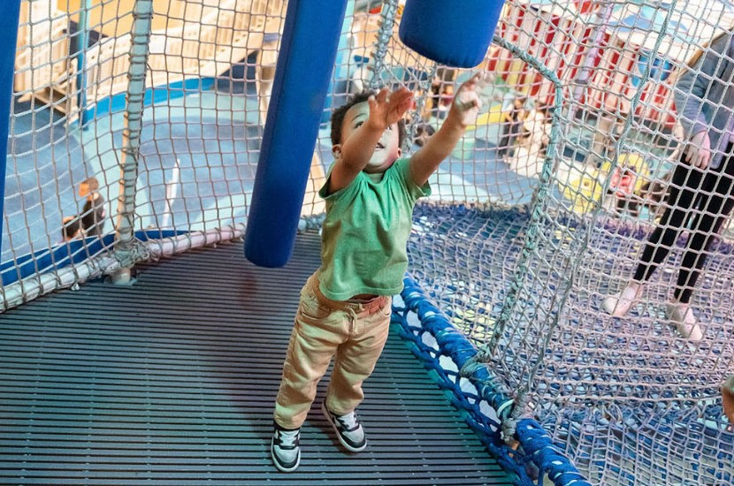 A young child standing and raising their arms up towards a soft, moveable blue bar in a netted play area