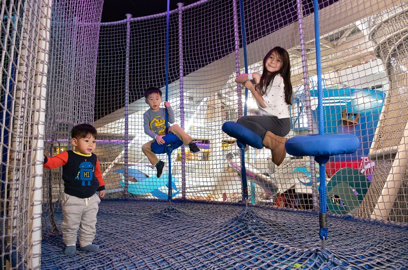 One children standing and two children sitting on rope swings in a netted play area overlooking some of Port Discovery's indoor exhibits