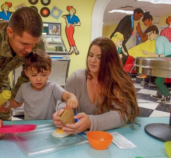 Two adults and a child playing together with pretend food and cookware at a table in Port Discovery's play diner