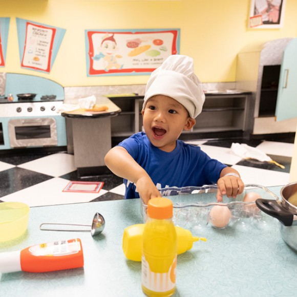 A young child wears a chef hat and pretends to mix ingredients in a pretend 50's style diner