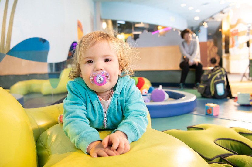A baby with a pacifier looking at the camera as they play and crawl on a soft green play mat in Port Discovery's Tot Trails exhibit