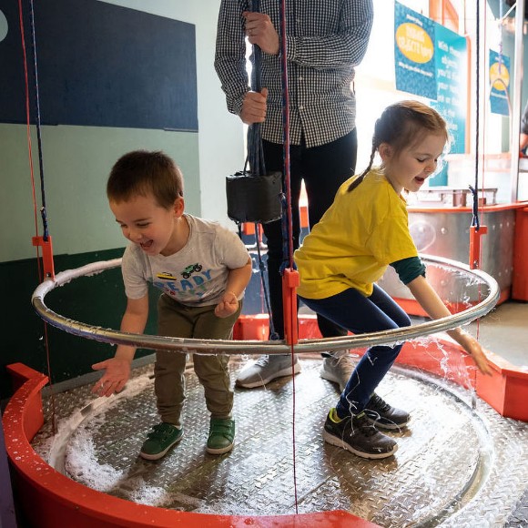 Two children smiling and laughing as they use their hands to pop a giant bubble that surrounds their bodies