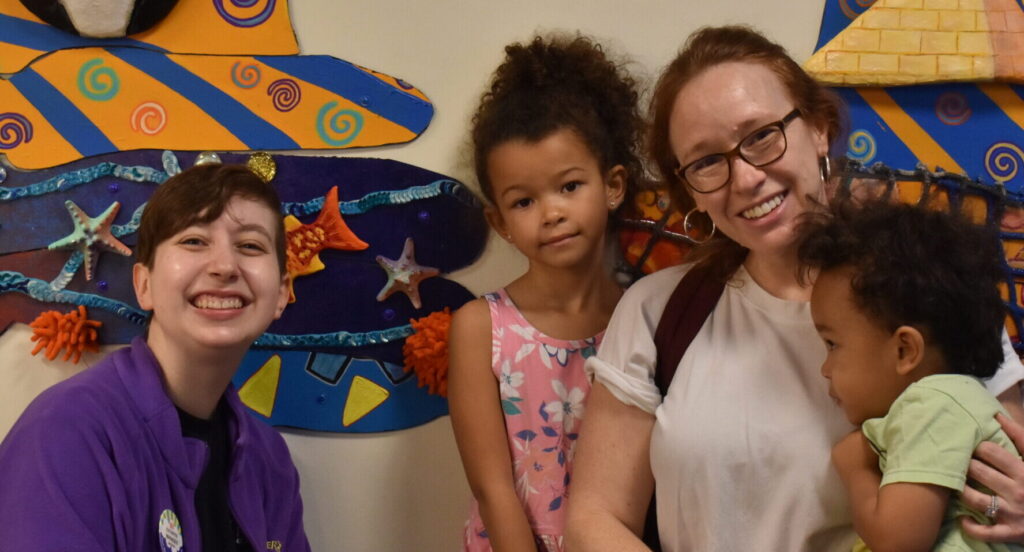 a Port Discovery employee standing next to a girl, a woman and a little boy in front of an artistic wall