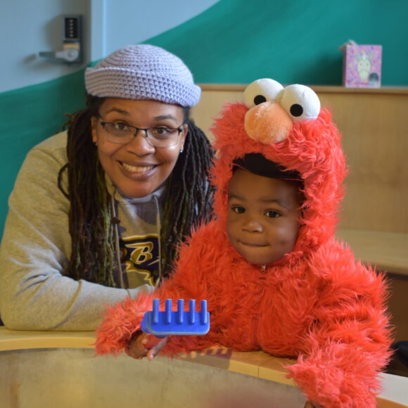 A child in an Elmo costume and their adult in front of a sandbox. 