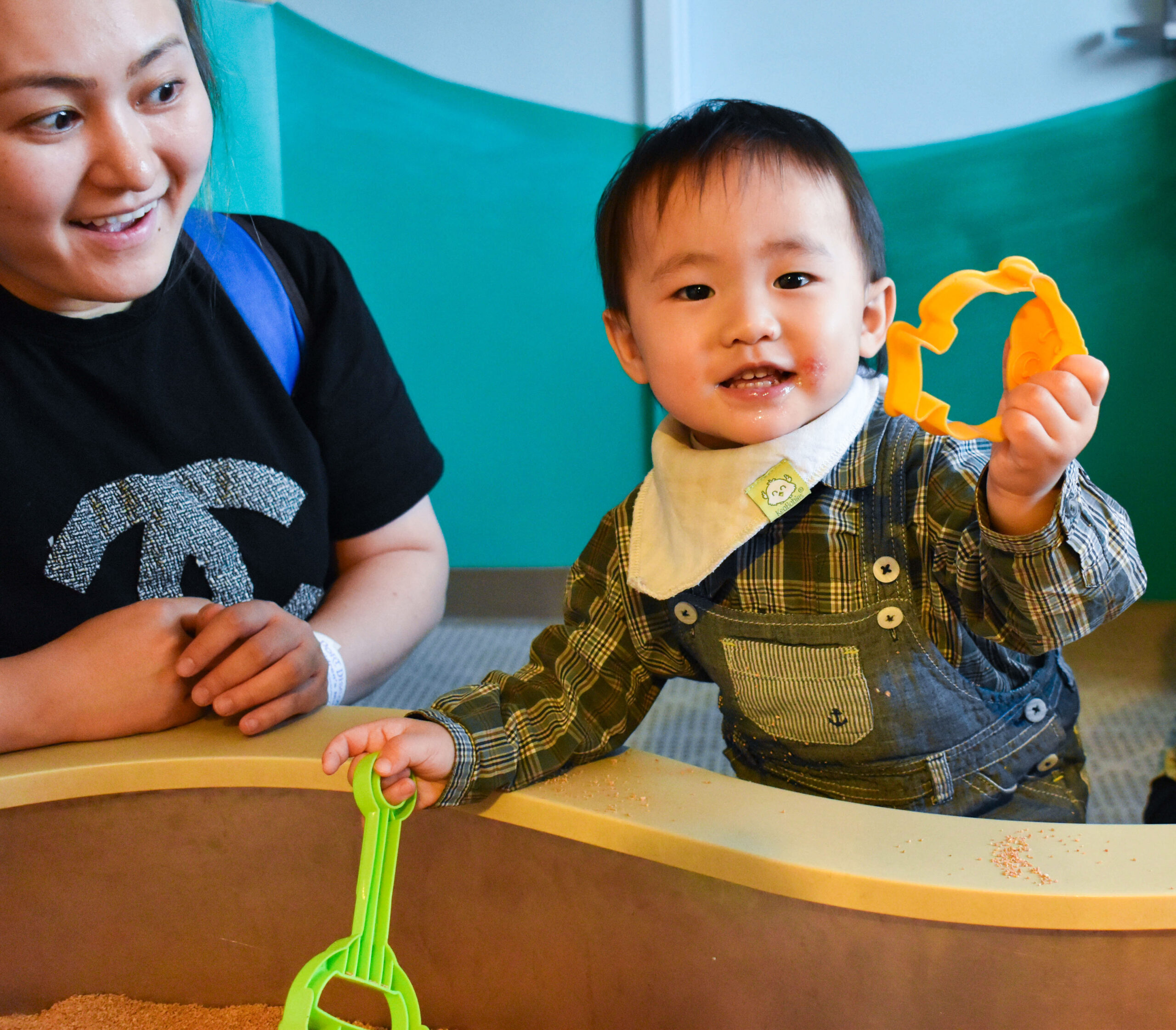 a toddler holding sandbox toy and smiling. Adult woman is smiling at the child.