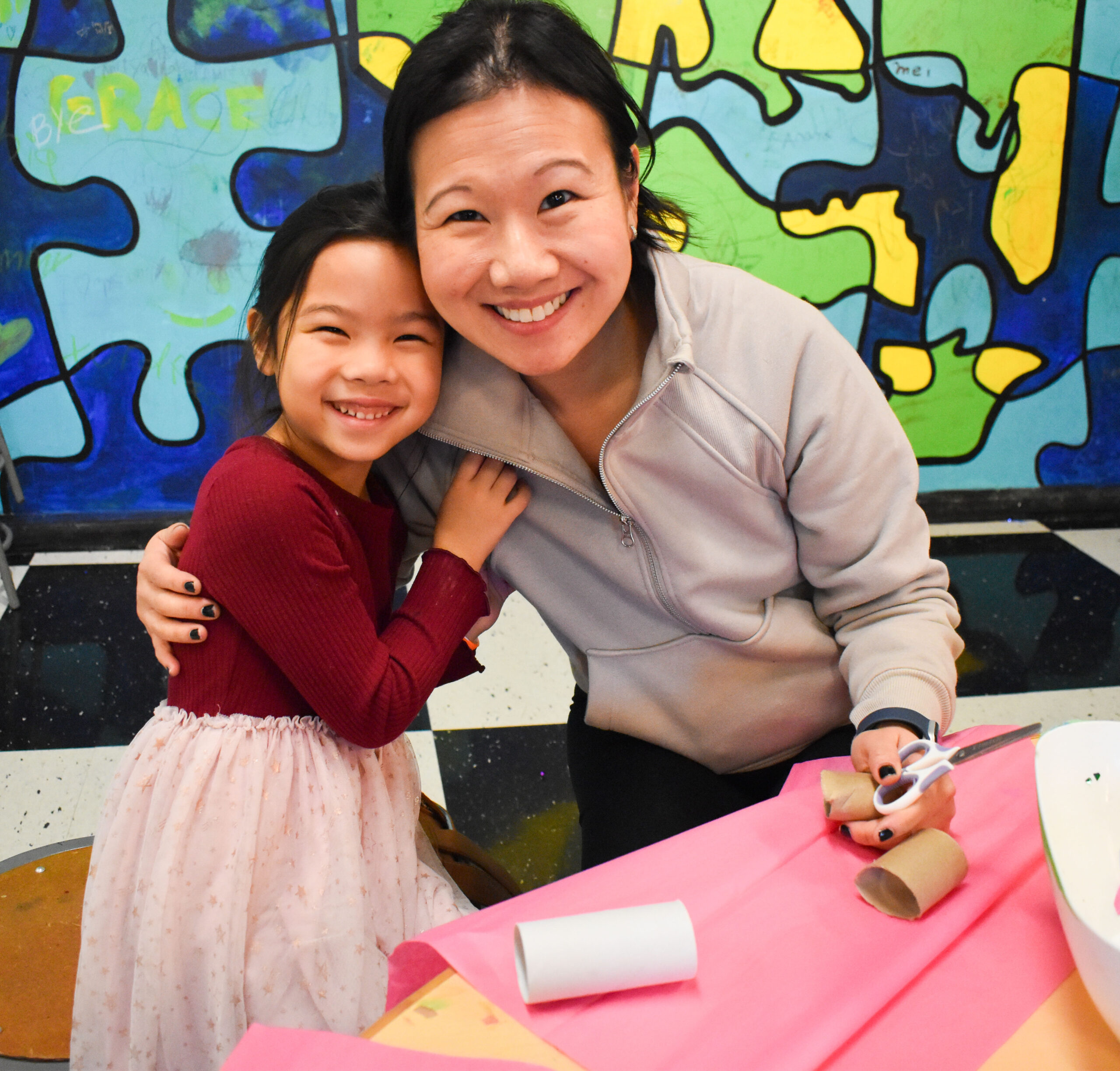 a woman and child sitting at a table and smiling with art supplies in front of them