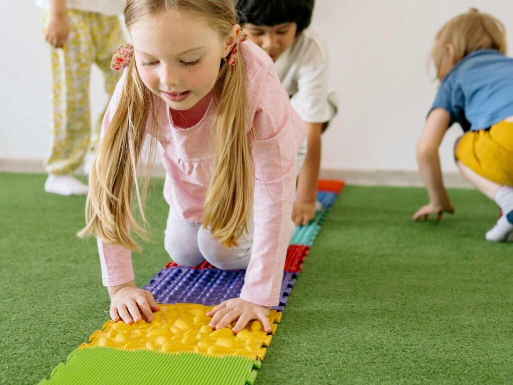 Four young children crawl across colorful textured sensory mats on green turf during a hands-on activity.