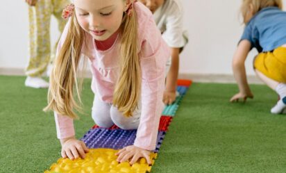 Four young children crawl across colorful textured sensory mats on green turf during a hands-on activity.