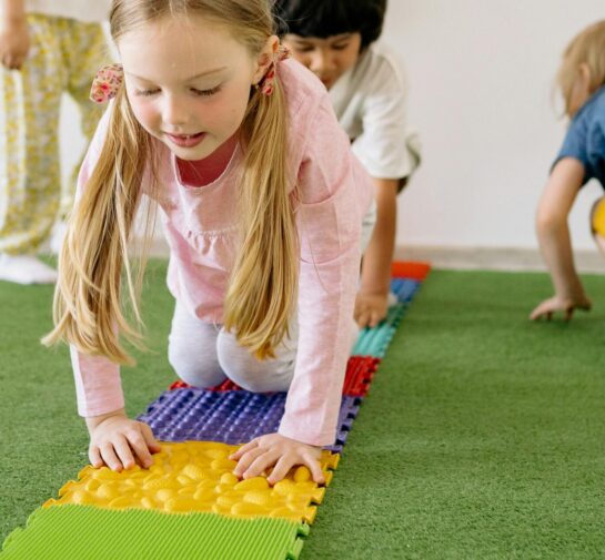 Four young children crawl across colorful textured sensory mats on green turf during a hands-on activity.